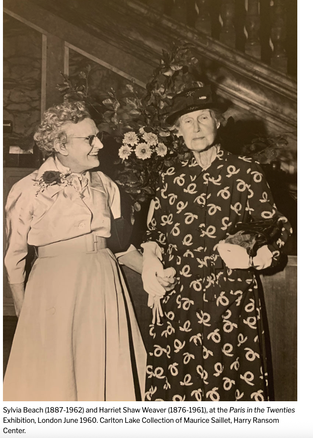 A 1960 photograph of Sylvia Beach and Harriet Shaw Weaver standing side by side at an exhibition in London, Beach laughing and Weaver in a patterned coat and hat.