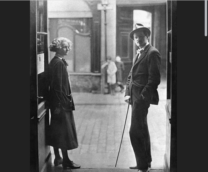 Sylvia Beach and James Joyce standing in the doorway of Shakespeare and Company in Paris, Beach turned toward the camera and Joyce in a hat and light suit leaning on a cane.