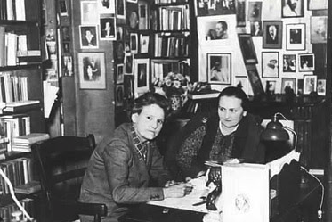 Sylvia Beach and Adrienne Monnier seated together at a desk in the bookshop, surrounded by bookshelves and a wall of framed author photographs.