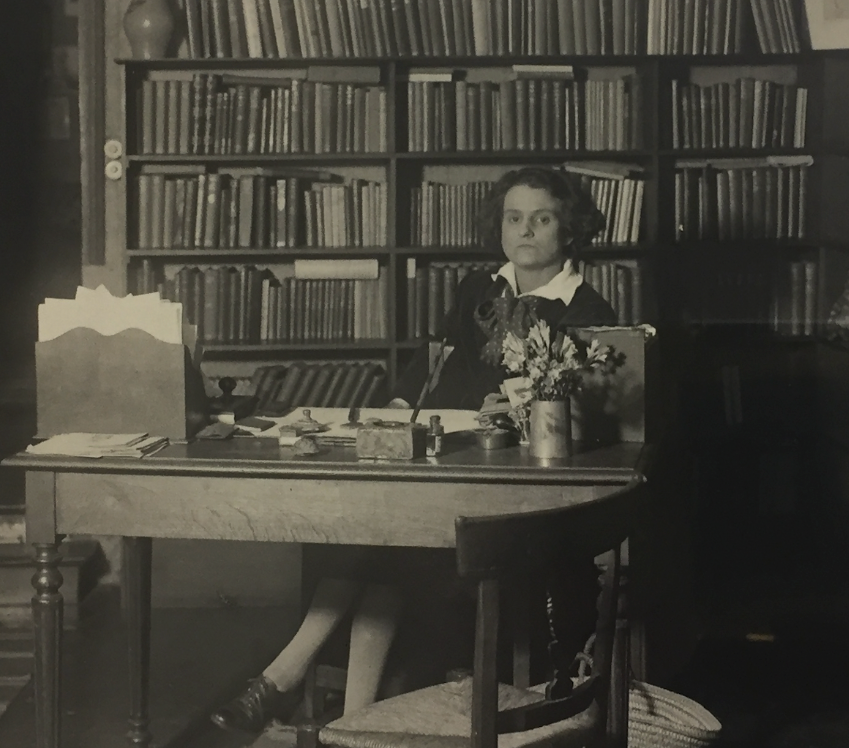 Sylvia Beach seated alone at a plain wooden desk in the shop, a vase of flowers and a letter rack in front of her and floor-to-ceiling bookshelves behind.