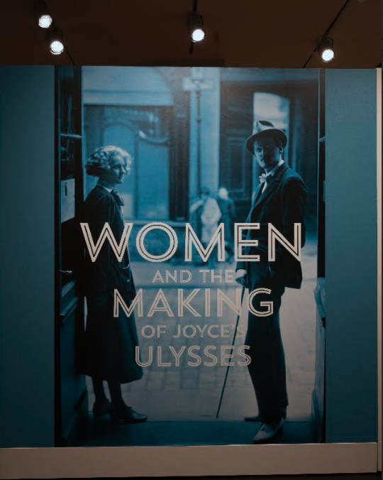 The title wall of the exhibition Women and the Making of Joyce's Ulysses, a blue-lit photographic mural of a woman and Joyce standing in a doorway with the exhibition title lettered across it.